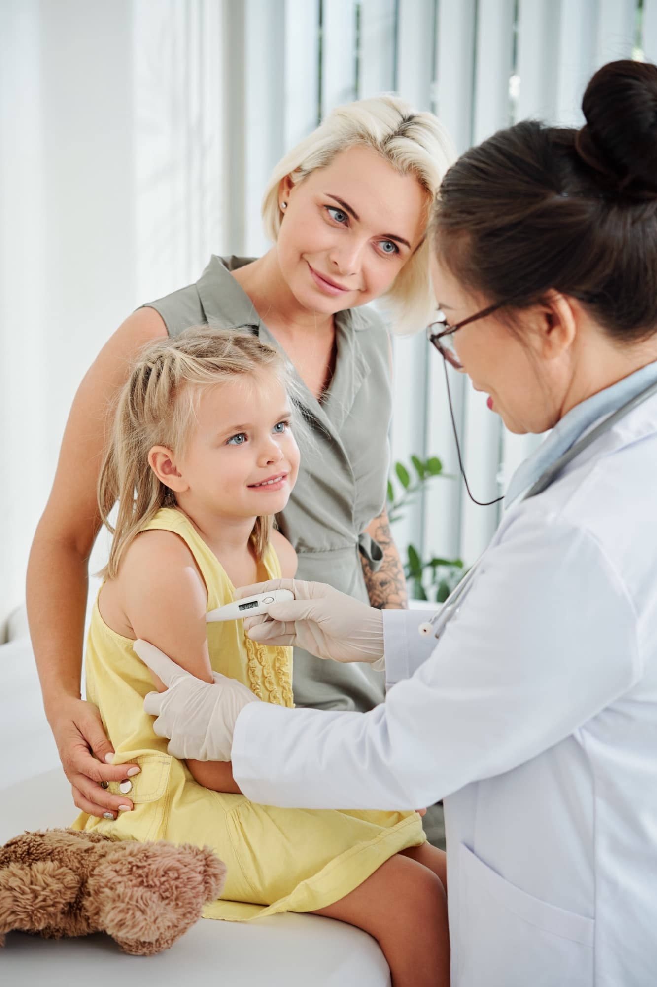 A pediatrician checking a young girl's temperature while her mother sits beside her